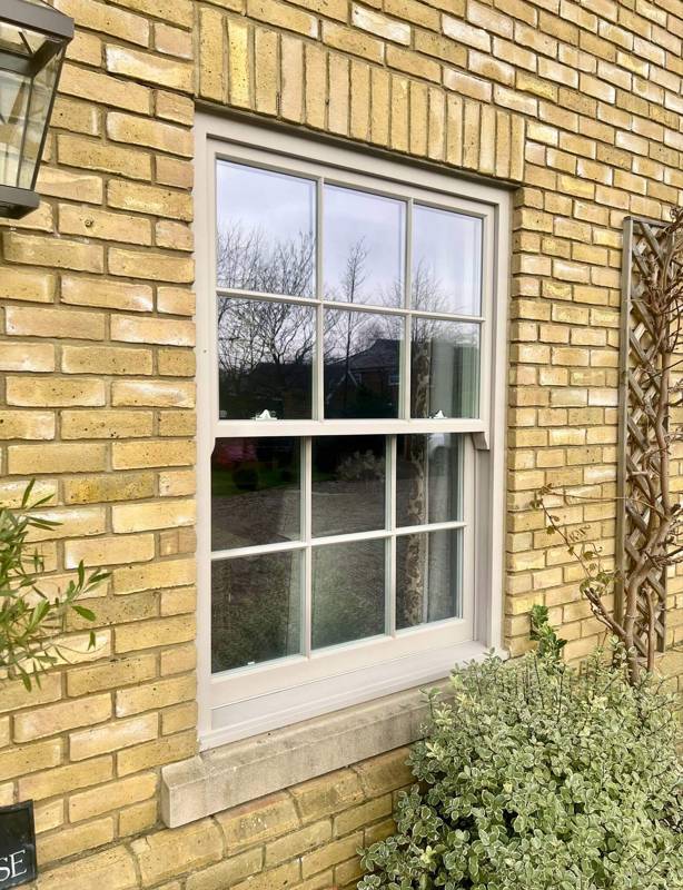 A double-hung sash window with a white frame set into a yellow brick wall. The window has six panes on each sash, and it reflects trees and sky from outside. A small bush is in front of the window, and a lantern-style light fixture is mounted on the wall to the left.