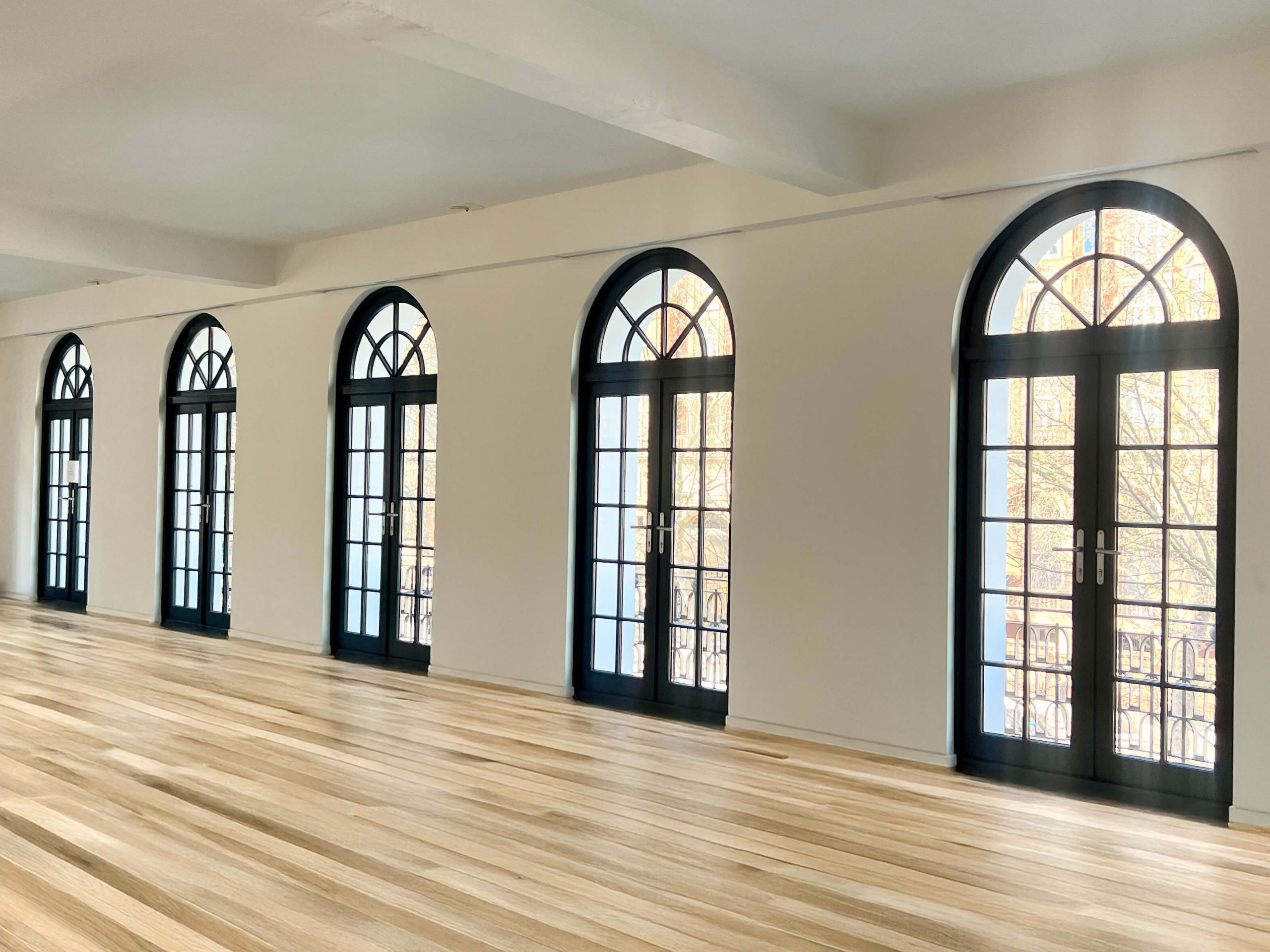 A spacious interior with elegant black-framed wooden French doors featuring semi-circular fanlights. The doors, arranged in a row, allow natural light to flood the room and offer access to a balcony with decorative wrought iron railings. The space is complemented by a warm-toned wooden floor, white walls, and a minimalist ceiling design, creating a sophisticated and airy atmosphere.