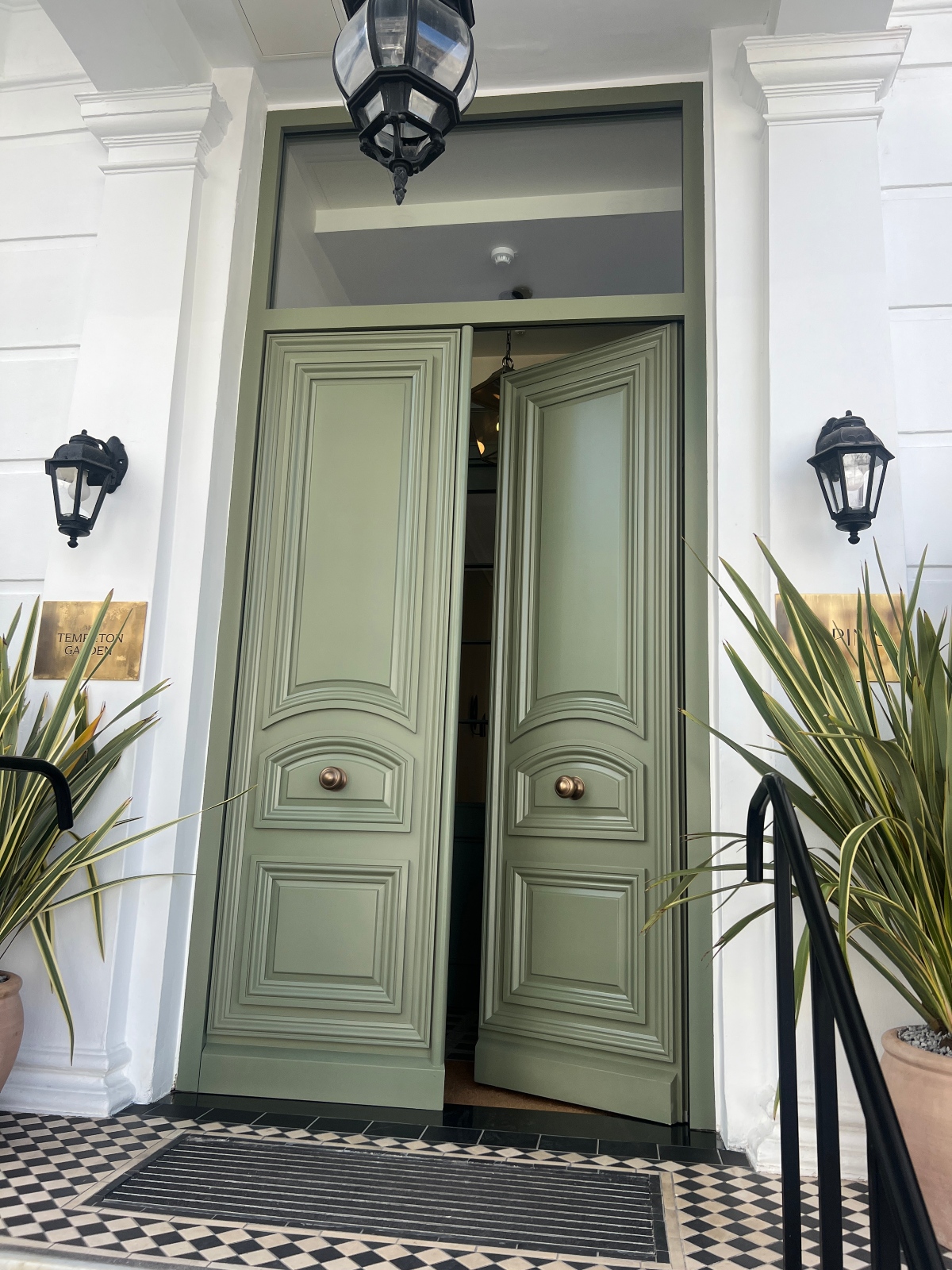 A set of grand, double timber doors, painted a muted green and featuring classical paneling, stand slightly ajar. These doors, made by Lomax + Wood, are set within a white facade with fluted pilasters. Two black lanterns adorn the wall on either side of the entrance, and potted plants sit on a checkered tile entryway.