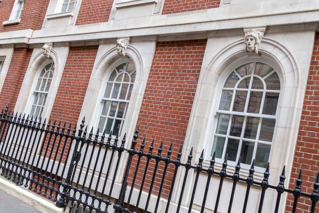 A row of bespoke arched timber sash windows set in a red brick building with white stone trim. Each window has a decorative keystone featuring a sculpted human face above it. The windows have white frames and are divided into smaller panes with a fanlight design at the top. A black wrought-iron fence with pointed finials runs along the sidewalk in front of the building. The reflection of nearby buildings is visible in the window glass.