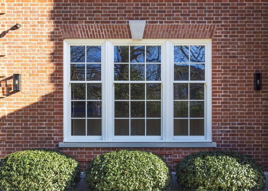 A close-up shot of a white timber box sash window with traditional crossbars, installed in a red brick wall. The window has a white sill and is flanked by three small bushes.