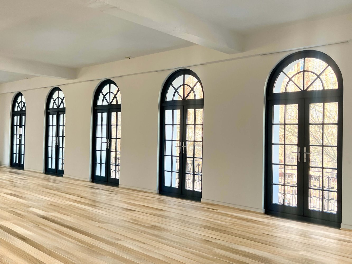 A spacious interior with elegant black-framed wooden French doors featuring semi-circular fanlights. The doors, arranged in a row, allow natural light to flood the room and offer access to a balcony with decorative wrought iron railings. The space is complemented by a warm-toned wooden floor, white walls, and a minimalist ceiling design, creating a sophisticated and airy atmosphere.