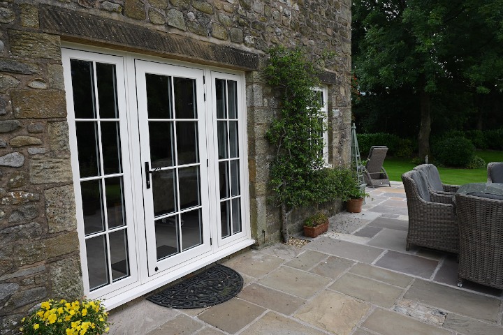 Showcasing new Residence Collection R9 and R7 doors from Lomax + Wood, featuring white frames and classic glazing bars on a stone building with a patio.