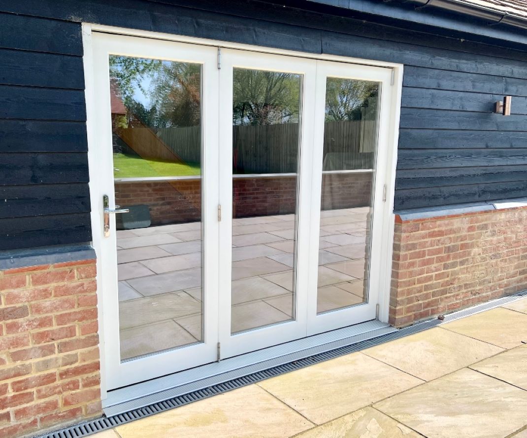 A bespoke, white timber bi-folding doorset with three leaves, set into a dark timber and red brick wall. The doors have large glazed panels, reflecting the patio and garden. A metal drainage channel runs along the base of the doors